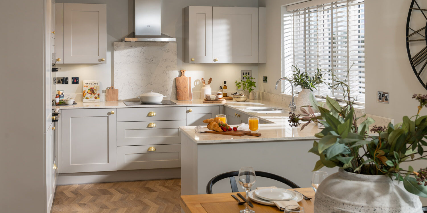 Kitchen with grey units and wooden flooring with table and chairs and vase with flowers at the forefront